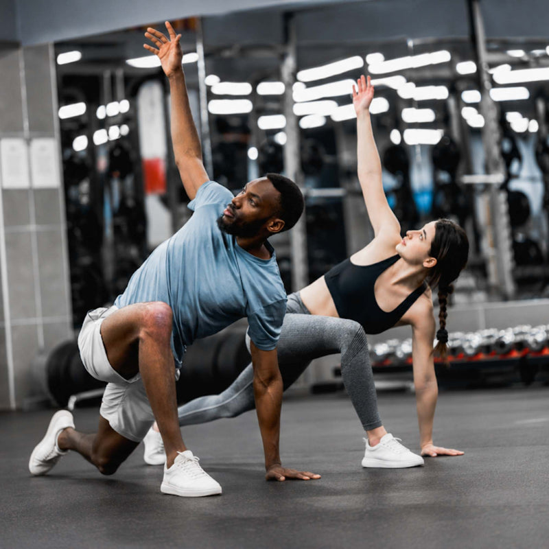Two people exercising in a gym, performing a side stretch pose on the floor. One person is wearing a light blue t-shirt, white shorts, and white sneakers, and the other is wearing a black sports bra, gray leggings, and white sneakers.