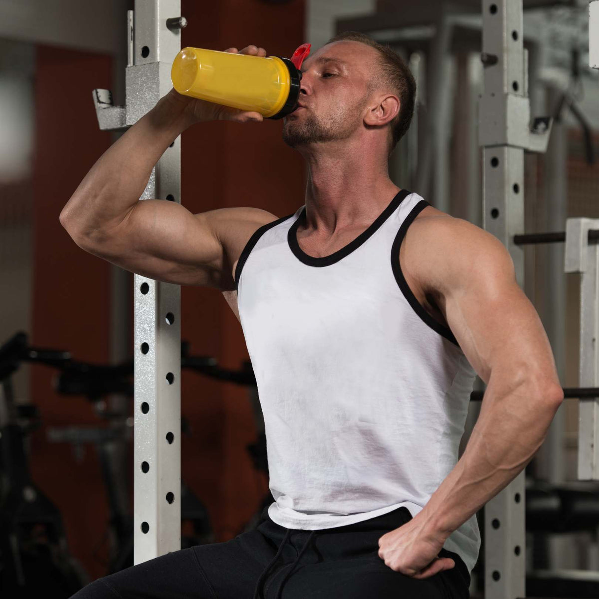 A muscular man in a gym drinking from a yellow shaker bottle while wearing a white tank top with black trim and black sweatpants.