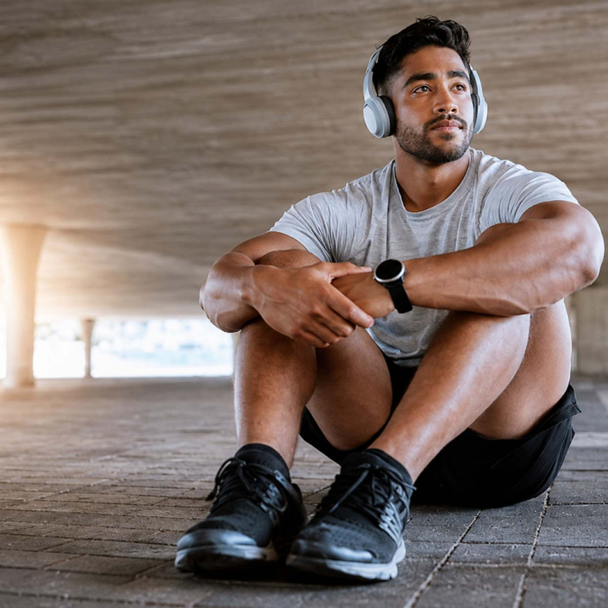 A man sitting on a concrete floor wearing white over-ear headphones, a light gray t-shirt, black shorts, and black athletic shoes, with a smartwatch on his wrist.