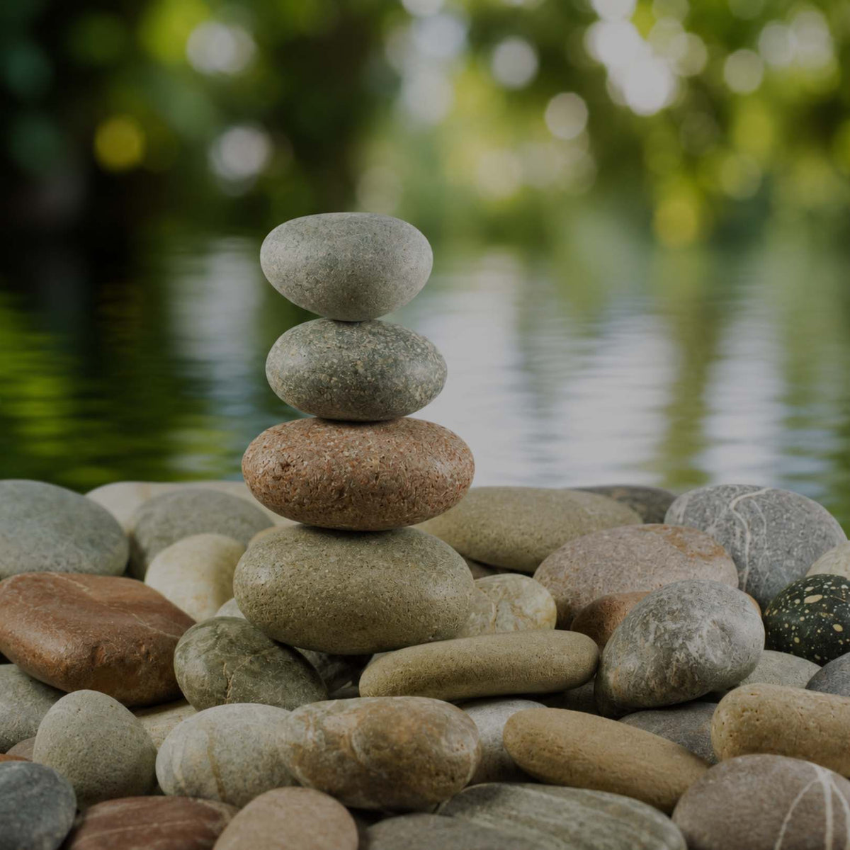 A stack of four carefully balanced smooth stones stands upright on a pile of similarly rounded stones, set against a softly blurred green and white water background.